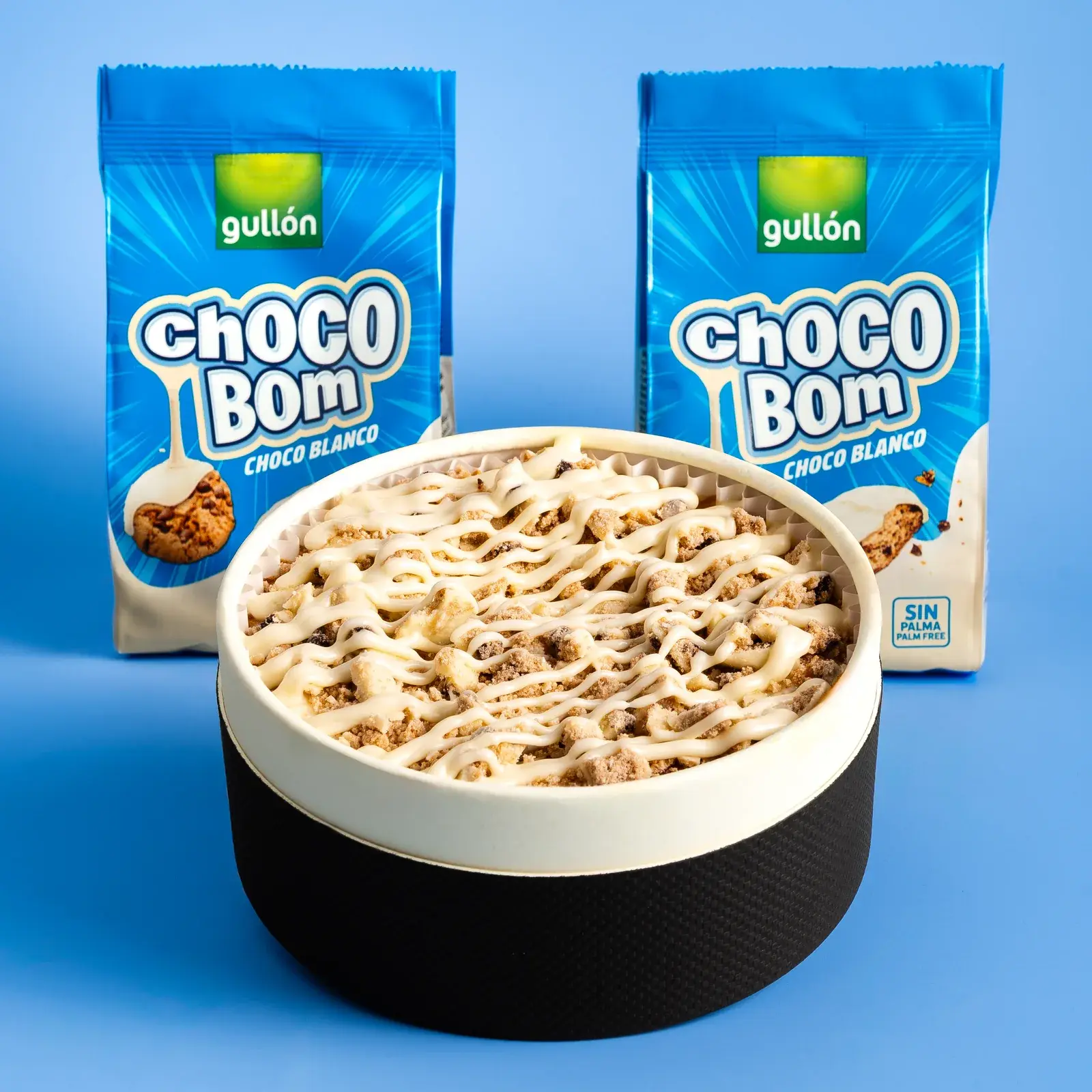 Tarta en su caja negra, cubierta de galleta troceada e hilos de chocolate blanco, delante de dos paquetes de galletas Chocobom Choco Blanco de Gullón sobre fondo azul